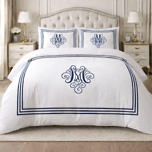 Può includere: Set di biancheria da letto bianco con finiture blu navy e dettagli ricamati. Il set comprende un copripiumino e due federe, ciascuna con un monogramma ornato. Il letto è appoggiato a una parete dai toni neutri, creando un'estetica classica ed elegante.