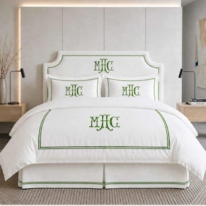 May include: A white bedding set featuring a green embroidered monogram with the letters MHC. The set includes a duvet cover, two pillowcases, and a bed skirt, all accented with a double green border. The monogram is centered on each piece.