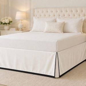 May include: A white bed skirt featuring a tailored design with crisp pleats and a contrasting black trim along the bottom edge. The fabric is smooth and opaque, designed to cover the base of the bed frame while providing a clean, structured look.