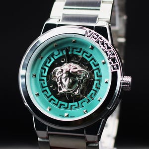 Op de afbeelding: Zilverkleurig Versace horloge met een turquoise wijzerplaat met het logo van het merk en een Medusa-hoofd. Het horloge heeft een zilveren band en een zwarte achtergrond. De wijzerplaat heeft een geometrisch ontwerp en kleine zilveren studs.