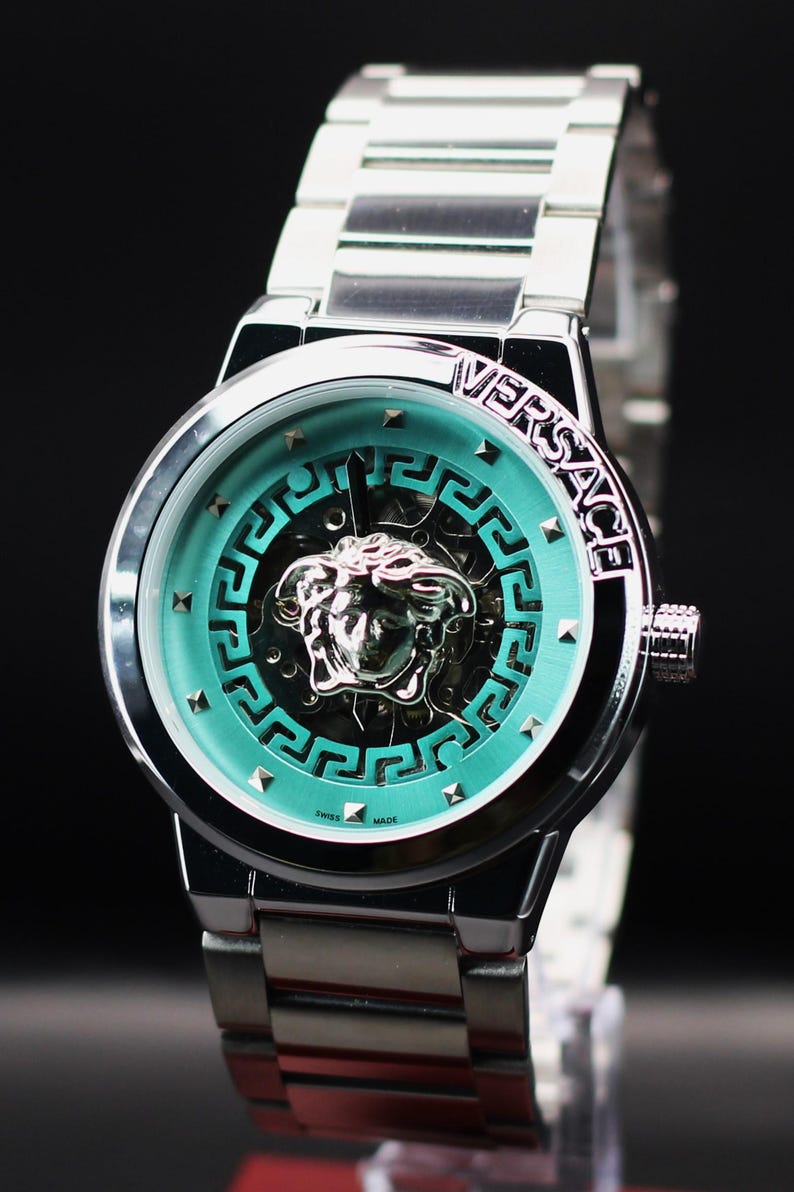 Op de afbeelding: Zilverkleurig Versace horloge met een turquoise wijzerplaat met het logo van het merk en een Medusa-hoofd. Het horloge heeft een zilveren band en een zwarte achtergrond. De wijzerplaat heeft een geometrisch ontwerp en kleine zilveren studs.