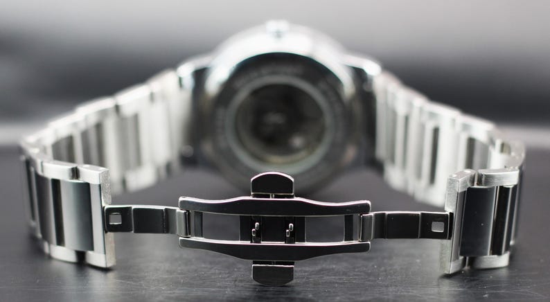 Op de afbeelding: Een close-up van een zilveren polshorloge met een roestvrijstalen band. De horlogeband is open en toont het sluitmechanisme. De wijzerplaat van het horloge is onscherp op de achtergrond.