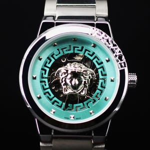 Op de afbeelding: Een zilverkleurig polshorloge met een turquoise wijzerplaat met een Medusa-hoofdontwerp. Het horloge heeft een zilveren band en het woord "VERSACE" is zichtbaar op de lunette. De wijzerplaat heeft een geometrisch patroon en de woorden "SWISS MADE".