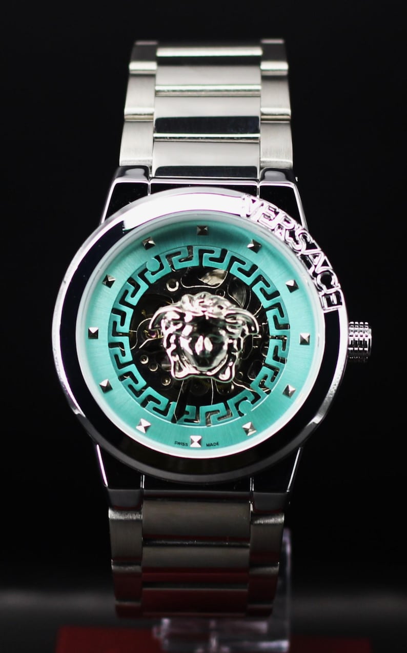 Op de afbeelding: Een zilverkleurig polshorloge met een turquoise wijzerplaat met een Medusa-hoofdontwerp. Het horloge heeft een zilveren band en het woord "VERSACE" is zichtbaar op de lunette. De wijzerplaat heeft een geometrisch patroon en de woorden "SWISS MADE".