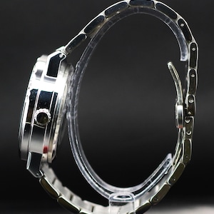 Op de afbeelding: Een zilverkleurig polshorloge met een metalen band. De wijzerplaat van het horloge is rond en heeft een donkere wijzerplaat. De band bestaat uit kleine, verbonden schakels. Het horloge wordt gepresenteerd op een transparante standaard tegen een donkere achtergrond.