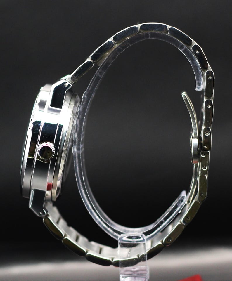 Op de afbeelding: Een zilverkleurig polshorloge met een metalen band. De wijzerplaat van het horloge is rond en heeft een donkere wijzerplaat. De band bestaat uit kleine, verbonden schakels. Het horloge wordt gepresenteerd op een transparante standaard tegen een donkere achtergrond.