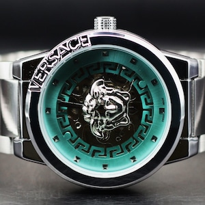 Op de afbeelding: Zilverkleurig Versace-horloge met een turquoise wijzerplaat en een zilveren Medusa-hoofd-embleem. Het horloge heeft een roestvrijstalen band en de merknaam staat boven de wijzerplaat.