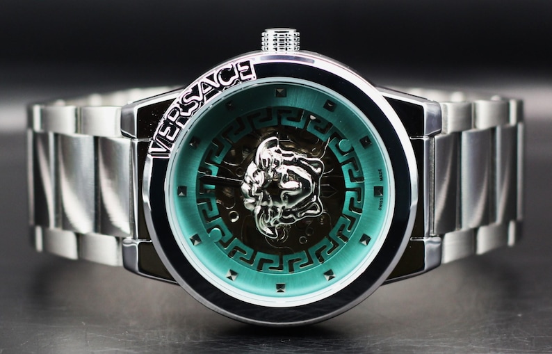 Op de afbeelding: Zilverkleurig Versace-horloge met een turquoise wijzerplaat en een zilveren Medusa-hoofd-embleem. Het horloge heeft een roestvrijstalen band en de merknaam staat boven de wijzerplaat.