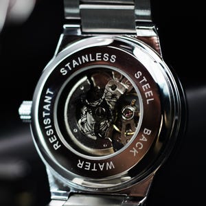 Op de afbeelding: Close-up van de achterkant van een roestvrijstalen horloge met zichtbare tandwielen. De achterkant van het horloge is gegraveerd met de woorden "STAINLESS STEEL BACK" en "WATER RESISTANT". De horlogeband is zilver.