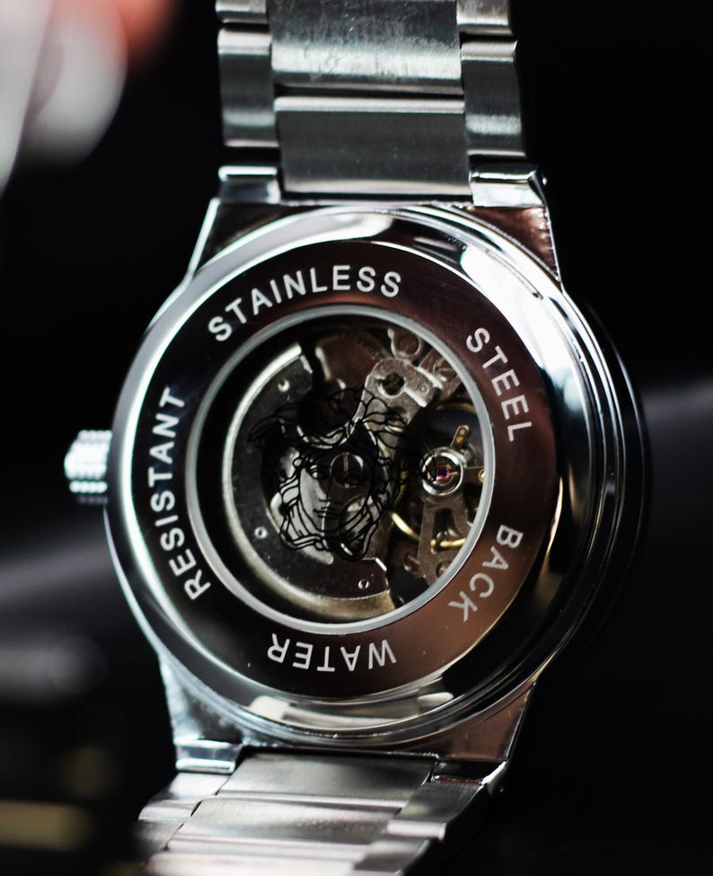 Op de afbeelding: Close-up van de achterkant van een roestvrijstalen horloge met zichtbare tandwielen. De achterkant van het horloge is gegraveerd met de woorden "STAINLESS STEEL BACK" en "WATER RESISTANT". De horlogeband is zilver.