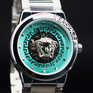 Op de afbeelding: Zilverkleurig polshorloge met een turquoise wijzerplaat met een Medusa-hoofd embleem en geometrisch ontwerp. Het horloge heeft een roestvrijstalen band en het woord "VERSACE" is zichtbaar op de bezel. Het horloge is gemarkeerd met "SWISS MADE".