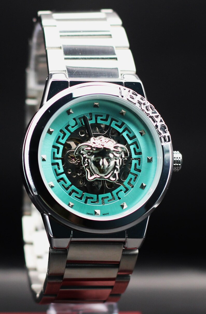 Op de afbeelding: Zilverkleurig polshorloge met een turquoise wijzerplaat met een Medusa-hoofd embleem en geometrisch ontwerp. Het horloge heeft een roestvrijstalen band en het woord "VERSACE" is zichtbaar op de bezel. Het horloge is gemarkeerd met "SWISS MADE".