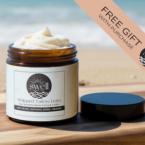 Op de afbeelding: Een potje opgeklopte talkbalsem met een zwarte deksel, op een houten oppervlak. De pot heeft een wit etiket met de tekst "Swell Beauty Co" en "Whipped Tallow Balm". Een banner leest "GRATIS CADEAU BIJ AANKOOP".