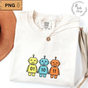 Peut inclure: T-shirt Comfort Colors crème avec trois robots de dessin animé jaunes, bleus et oranges. Chaque robot a une expression différente et un numéro : 01, 10 et 11. Un collier en or avec un pendentif est drapé sur le t-shirt.