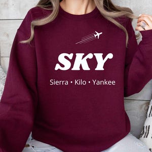May include: Burgundy sweatshirt with the word "SKY" in white, a small airplane graphic, and the text "Sierra • Kilo • Yankee." The sweatshirt is a solid color and has a crew neck.
