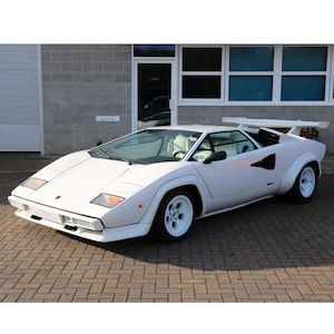Pode incluir: Um carro esportivo Lamborghini Countach branco com um design em forma de cunha distinto. O carro apresenta uma asa traseira, rodas brancas e um perfil baixo. O veículo está estacionado em uma superfície de tijolo em frente a um edifício.