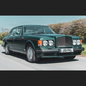 Pode incluir: Um sedã vintage verde escuro com uma grelha cromada e faróis, dirigindo em uma estrada pavimentada. O carro tem uma placa de licença e está contra um fundo desfocado de árvores e céu azul.
