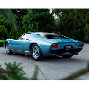 May include: A metallic light blue sports car with a low-profile body, silver wheels, and black rear window louvers. The vehicle features a black license plate with the text P400S, rectangular red taillights, and a dark rear grille.