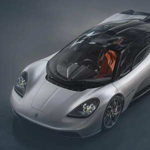 以下が含まれることがあります： 黒い屋根とオレンジ色の内装のシルバーのスポーツカー。 大きなヘッドライトと低いプロファイルを備えた、洗練された空力設計の車です。 ホイールは黒とシルバーのアクセントです。 車は灰色の表面に駐車されています。