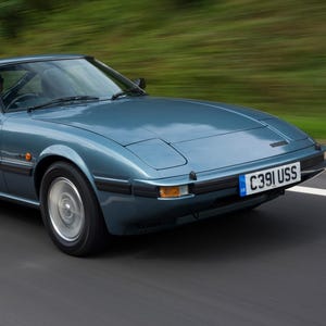 Pode incluir: Um carro esportivo vintage azul-petróleo acelera em uma rodovia. O carro tem um design elegante e aerodinâmico, com faróis retangulares e uma placa que diz "C391 USS". O fundo está desfocado, sugerindo movimento.
