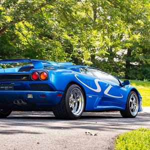Pode incluir: Um carro esportivo Lamborghini Diablo azul com listras brancas. O carro tem um spoiler traseiro, rodas cromadas e lanternas traseiras vermelhas. O veículo está estacionado em uma estrada pavimentada com uma área gramada verde e árvores ao fundo.