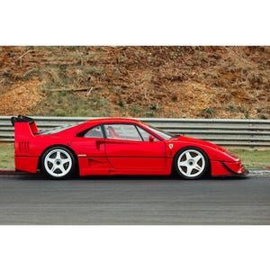 Pode incluir: Um carro esportivo Ferrari vermelho com rodas brancas está estacionado em uma pista de corrida. O carro tem um spoiler e um perfil baixo. O fundo apresenta uma colina gramada e uma barreira branca e cinza.
