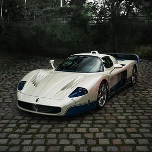May include: A white Maserati MC12 sports car with blue lower body accents, a low-profile aerodynamic chassis, a prominent front grille featuring the trident emblem, and a rear wing, parked on a cobblestone surface.
