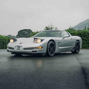 May include: A silver Chevrolet Corvette sports car with pop-up headlights illuminated, black multi-spoke wheels, and a front license plate reading X114 UAO. The vehicle is positioned on a dark asphalt surface under an overcast sky.