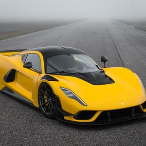 May include: A bright yellow sports car with a black roof, black side mirrors, and black wheels. The vehicle features a low-profile aerodynamic body, sharp angular headlights, and a black front splitter, positioned on a gray asphalt track.