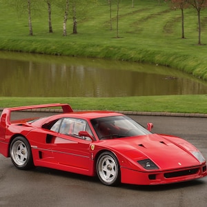 May include: A bright red Ferrari F40 sports car featuring a low-profile aerodynamic body, a large rear wing, pop-up headlights, and silver five-spoke wheels. The vehicle displays the iconic yellow Ferrari emblem on the side panel near the door.