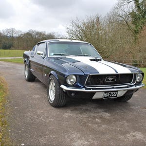 May include: A dark blue vintage Ford Mustang fastback with a prominent white racing stripe running down the center of the hood and roof. The car features chrome bumpers, round headlights, a black front grille, and a license plate reading MUF755F.