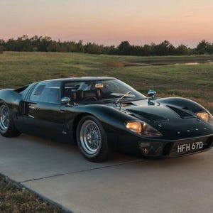 May include: A dark green Ford GT40 sports car with a low-profile aerodynamic body, wire-spoke wheels, and round headlights. The vehicle features a front license plate reading HFH 67D and is positioned on a paved road during the golden hour.