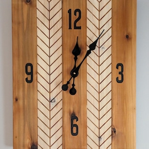 May include: Square wooden wall clock with a natural wood grain finish. The clock face features black numerals at 3, 6, 9, and 12 o'clock, with black clock hands. The center panel has a chevron pattern.