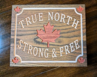 Może przedstawiać: Drewniany znak z wygrawerowanymi słowami "TRUE NORTH STRONG & FREE". Znak posiada czerwony liść klonu pośrodku i mniejsze akcenty liści klonu w każdym rogu. Znak ma białą obwódkę.