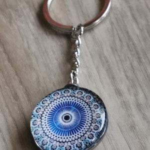 Peut inclure: Un porte-clés argenté avec un pendentif rond orné d'un motif géométrique bleu et blanc. Le pendentif a une finition semblable à du verre, et le porte-clés comprend un anneau fendu et une chaîne décorative.