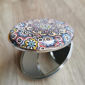 Puede incluir: Un espejo compacto redondo plateado con un colorido e intrincado diseño de mosaico en la parte superior. El mosaico presenta una variedad de colores, incluyendo rojo, azul, amarillo y blanco. La palabra "MOROCCO" está impresa en la parte superior. El espejo está abierto.