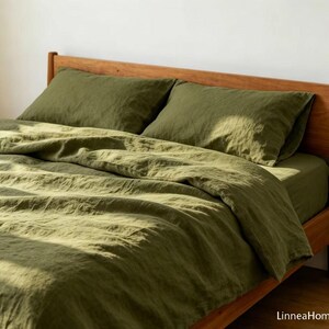 May include: A bed set featuring an olive green duvet cover and two matching pillowcases with a visible textured fabric weave. The bedding is arranged on a wooden bed frame, with the text LinneaHome appearing in the bottom right corner.