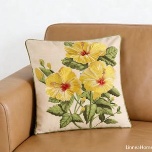May include: Square decorative pillow with embroidered yellow hibiscus flowers, red centers, and green leaves. The pillow has a cream-colored background with a green border. The pillow is on a brown leather couch.