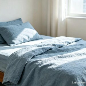 May include: A dusty blue bedding set consisting of a duvet cover and two matching pillowcases. The fabric has a visible textured weave and is draped over a bed. The text LinneaHome is printed in the bottom right corner of the image.