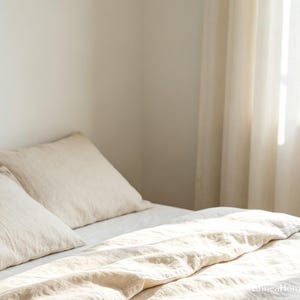 May include: A bed featuring a light beige duvet cover and two matching pillows. The bedding has a visible woven texture and is arranged neatly. Natural light streams in from the side, illuminating the fabric and creating soft shadows across the surface.