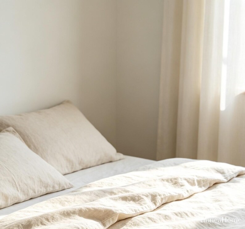 May include: A bed featuring a light beige duvet cover and two matching pillows. The bedding has a visible woven texture and is arranged neatly. Natural light streams in from the side, illuminating the fabric and creating soft shadows across the surface.