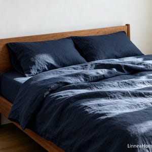 May include: A dark navy blue bedding set featuring a duvet cover and two matching pillowcases with a visible textured fabric weave. The bedding is arranged on a wooden bed frame, with the text LinneaHome visible in the bottom right corner.