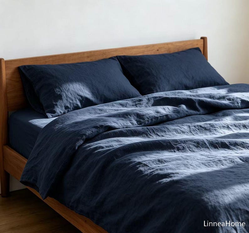 May include: A dark navy blue bedding set featuring a duvet cover and two matching pillowcases with a visible textured fabric weave. The bedding is arranged on a wooden bed frame, with the text LinneaHome visible in the bottom right corner.