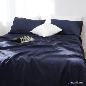 May include: A navy blue bed set with a duvet cover and pillowcases. The bed is made with a dark blue sheet, two navy blue pillowcases, and two white pillows. A small bottle and sprig of greenery are on the bed.