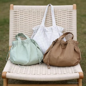 May include: Three fabric tote bags and drawstring bags in various colours sit on a woven chair. The bags include a white grid pattern tote bag, a light green drawstring bag, and a brown drawstring bag. The bags have handles.
