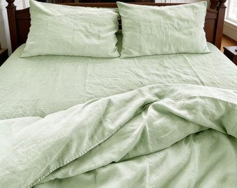 May include: A light sage green bedding set featuring a flat sheet, a duvet cover, and two matching rectangular pillowcases. The fabric has a visible, slightly textured weave and a soft, rumpled appearance. The brand name LinneaHome is visible.