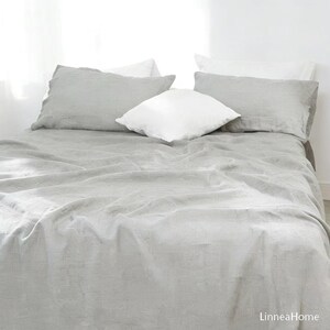 May include: A light gray linen bed sheet covering a mattress, accompanied by two matching light gray rectangular pillowcases and two white square pillows. The text LinneaHome is visible in the bottom right corner of the image.