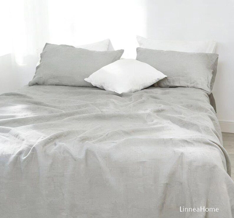 May include: A light gray linen bed sheet covering a mattress, accompanied by two matching light gray rectangular pillowcases and two white square pillows. The text LinneaHome is visible in the bottom right corner of the image.