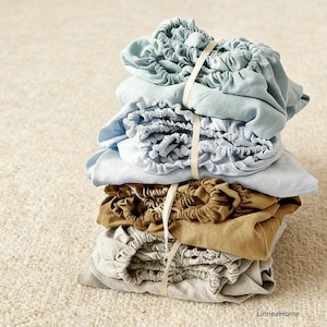 Peut inclure: Une pile de draps de lit pliés de différentes couleurs, dont bleu clair, beige et gris, attachés avec un ruban crème. Les draps semblent être faits d'un tissu doux et texturé, soigneusement rangés.