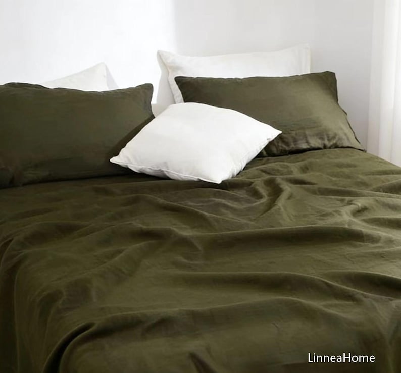 May include: A bed set featuring a dark olive green flat sheet and two matching rectangular pillowcases, complemented by two white square throw pillows. The fabric has a visible texture and the text LinneaHome appears in the bottom right corner.